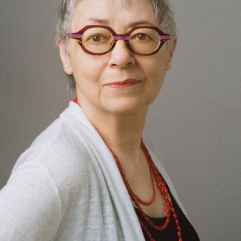 A person posed against a neutral gray background, looking over their shoulder. They have a short, textured haircut and are wearing vibrant, architectural eyewear.