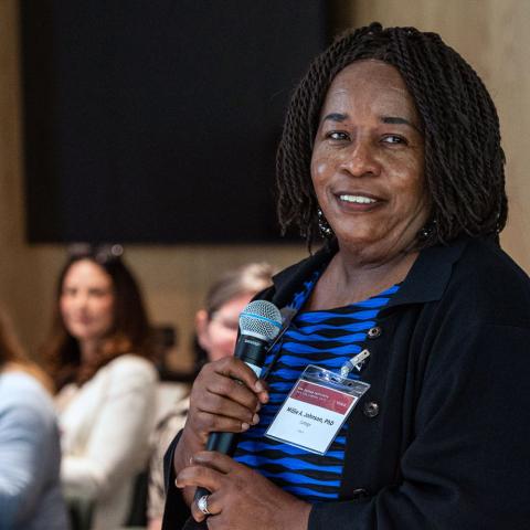 Person standing and speaking amid a seated smiling crowd.