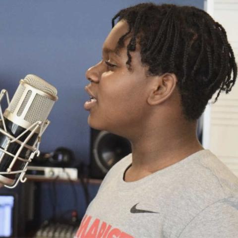 A child with curly black hair in braids speaks into a microphone.