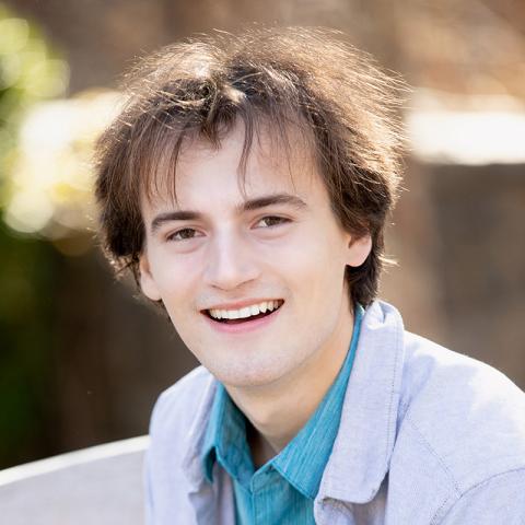 A person with brown hair smiling brightly while sitting outdoors on a wooden bench.