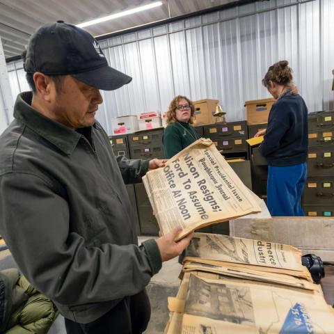 Person standing in an archive room holding and old yellowed newspaper with the front page showing.