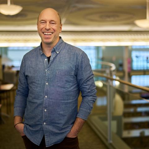 A smiling person wearing a denim shirt stands with their hands in their pants pockets in an indoor atrium with big windows.