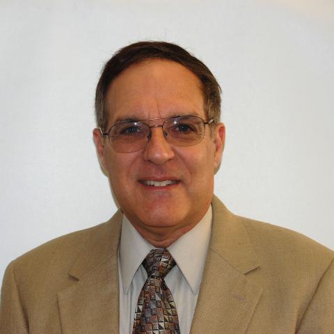 David Engel wearing glasses, a tan blazer, light-colored dress shirt, and patterned tie, smiling in front of a plain white background.