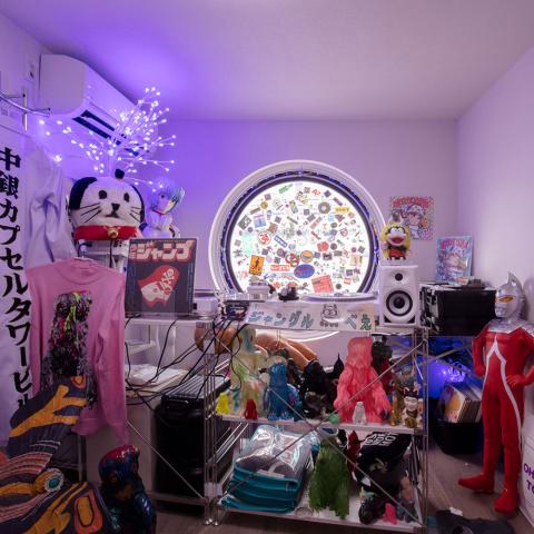 A compact, capsule-like room filled with colorful Japanese pop culture objects and media equipment, including a circular window covered with stickers, shelves of vinyl records and figurines, speakers and turntables, hanging garments with Japanese text, and a red Ultraman figure standing on the floor, illuminated by purple accent lighting.