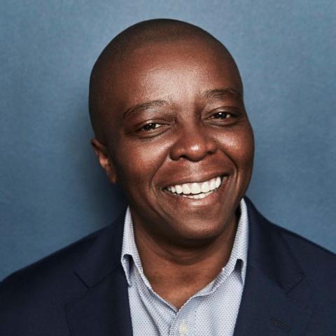 Head-and-shoulders portrait of filmmaker Yance Ford smiling warmly, wearing a navy blazer and light patterned shirt against a blue studio backdrop.