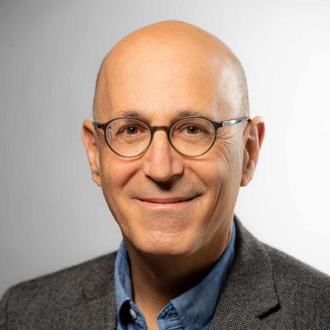 A bald individual with round glasses and a slight smile faces forward. They wear a light blue button-down shirt underneath a grey herringbone textured blazer. The background is a solid, neutral light grey.