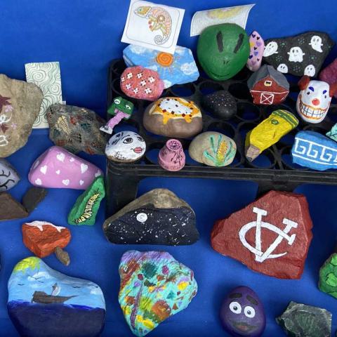 A collection of uniquely painted rocks is displayed against a blue background. The rocks showcase a variety of designs, including colorful patterns, whimsical creatures, and abstract motifs. A few notable pieces depict a ship, a chameleon, and a clown face, while others are adorned with hearts and spirals.
