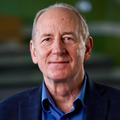 A person with light-colored, thinning hair looks toward the camera with a slight smile. The individual wears a blue button-down shirt under a dark, subtly patterned blazer. The background is softly blurred with shades of green and dark gray.