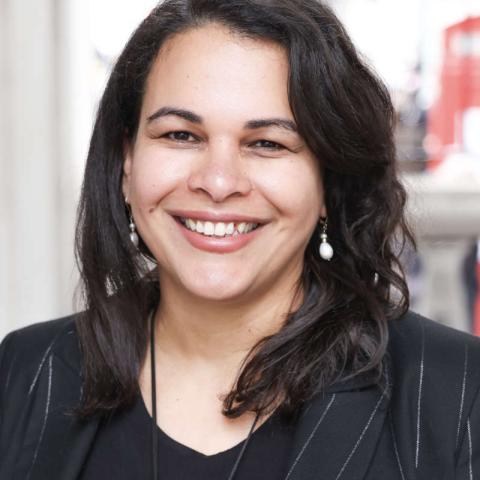 A photo of Rosa Andújar. They are smiling and wearing a dark blazer with subtle pinstripes and a black top.