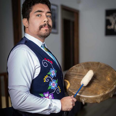 An individual stands holding a circular hand drum and a mallet with a soft white head. They wear a light-colored long-sleeve shirt under a navy vest decorated with colorful floral embroidery. The setting appears to be an indoor hallway.