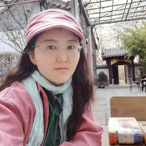 A figure is seated at a wooden table, wearing a pink cap and a light blue scarf over a layered outfit that includes a pink jacket and a green shirt. The figure is positioned slightly to the left of the frame, with arms crossed on the table. In the background, a wooden trellis can be seen above, partially obscured by bare branches, while hints of traditional architecture peek through, including a doorway framed in red. Soft, natural light illuminates the scene, creating a calm atmosphere.