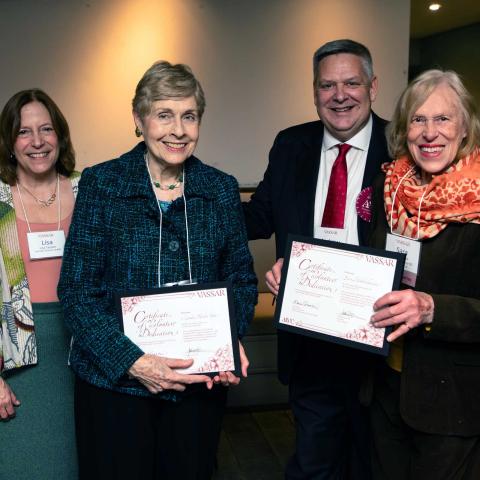 Four smiling people standing side by side while two in the center hold framed documents.