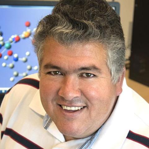A headshot of a smiling person with short gray hair, wearing a white polo shirt, with a molecular model display visible in the background.