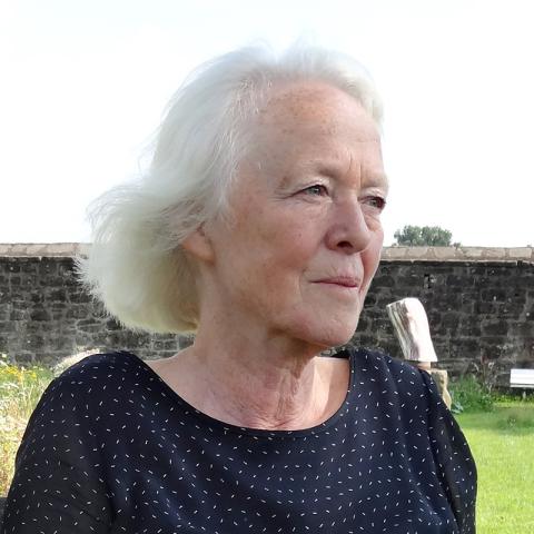Portrait of Barbara Beisinghoff seated outdoors in a garden, wearing a dark patterned top, with flowers, grass, and a stone wall in the background.