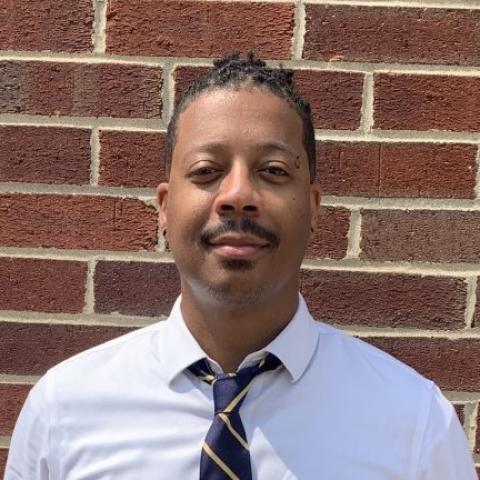 Head-and-shoulders portrait of Brandon A. Jackson standing in front of a brick wall, wearing a light-colored shirt and striped tie.