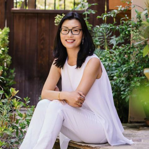 An individual with dark, shoulder-length hair and glasses smiles while sitting outdoors. They wear a white sleeveless top and white pants. Lush green plants and a wooden gate are in the background.