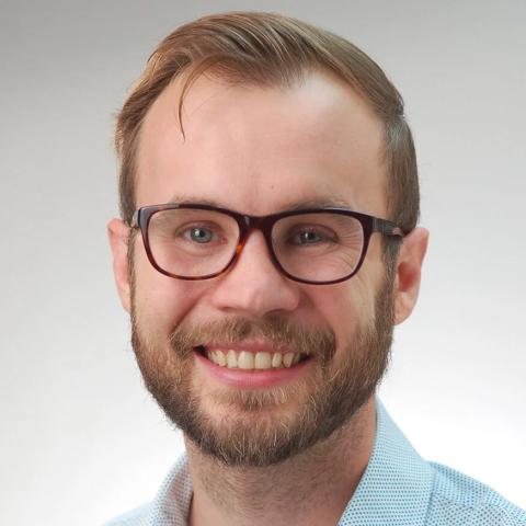 A headshot of a smiling person with short blond hair and a beard, wearing dark-rimmed glasses and a light blue collared shirt, against a neutral light gray background.
