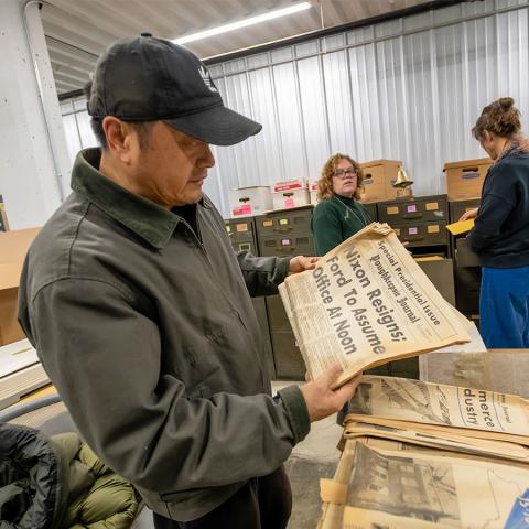 A person wearing a zip-front jacket and baseball cap holds an old issue of the Poughkeepsie Journal that has the headline "Nixon Resigns; Ford to Assume Office at Noon."