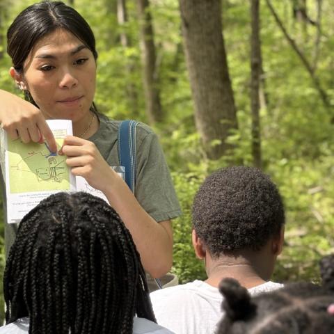 An individual stands in a wooded area, holding up a map and pointing to a specific trail location while addressing a group. The person wears a green shirt and a shoulder bag. The group, seen from behind, listens in a forest setting with green foliage.
