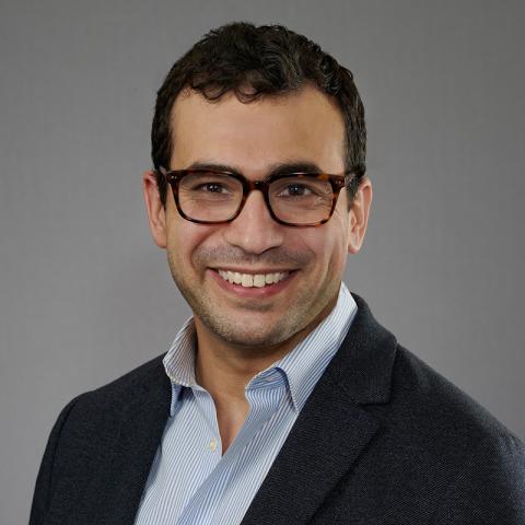 A headshot of a smiling person with short dark hair and tortoiseshell glasses, wearing a dark blazer over a blue striped shirt, against a neutral gray background.
