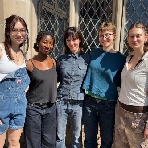Five students stand together outdoors in front of a brick building with leaded glass windows, smiling, with their arms around each other.