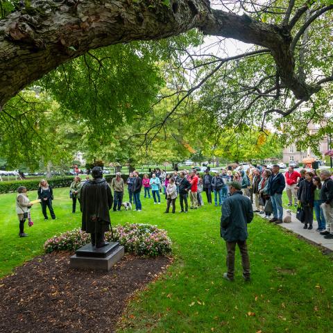 Group of visitors gathered on a campus lawn around a statue during an outdoor tour, with large tree branches overhead.