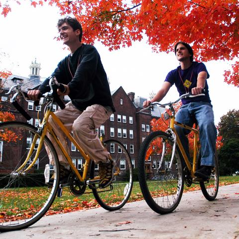 Two people riding bicycles along a campus path lined with vibrant red autumn leaves, with academic buildings in the background.