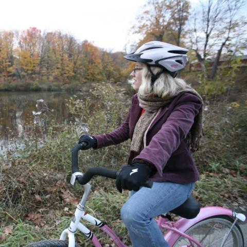 A person sits on a pink bicycle beside a calm body of water, framed by autumn foliage. The cyclist wears a white helmet and a purple jacket layered over a red shirt, with a brown scarf draped around their neck. In the background, trees display vibrant fall colors, casting reflections on the water's surface. The scene is set under a cloudy sky, suggesting a cool, overcast day, with earthy tones in the grass and fallen leaves enriching the natural landscape.