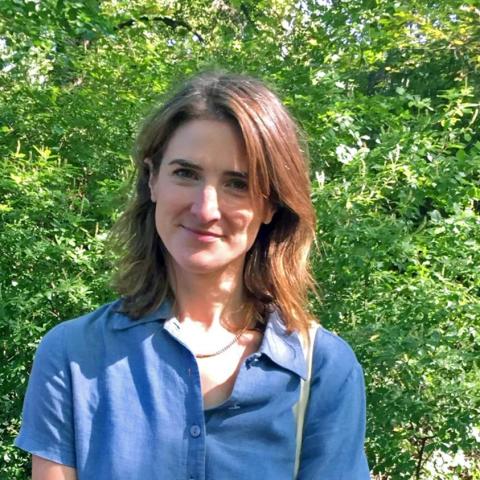 An individual with shoulder-length brown hair smiles while standing outdoors. They are wearing a blue short-sleeved button-down shirt and a thin gold necklace. Lush green foliage and sunlight-dappled leaves serve as the background.