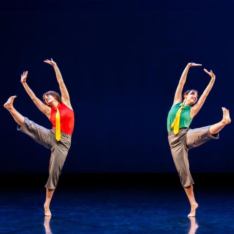 Two individuals on a stage perform a synchronized dance move against a dark blue backdrop. Each person balances on one leg with the other extended outward, wearing grey trousers, a yellow tie, and a sleeveless shirt—one in red and one in green.