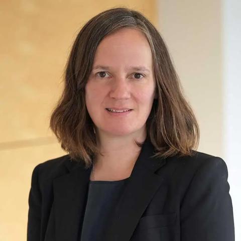 Professional headshot of a person with shoulder-length brown hair, wearing a black blazer and top, with a slight smile against a soft, neutral background.
