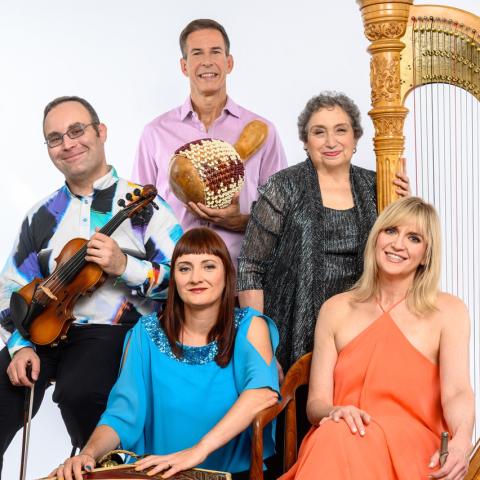 Group portrait of five musicians posing with their instruments against a light gray background. One person holds a violin, another stands holding a beaded gourd percussion instrument, and a large harp is visible at the right side of the image. Two seated performers wear bright blue and coral outfits, while the standing performers wear patterned and metallic attire. The group smiles toward the camera in a formal promotional-style photo.