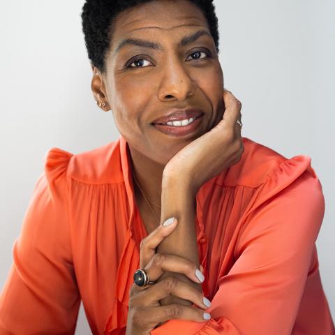 Ayana Mathis with short natural hair wearing a coral blouse, resting their chin on one hand and smiling against a light gray background.