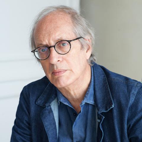 Portrait of a person with glasses wearing a blue shirt and jacket, seated indoors against a neutral background.