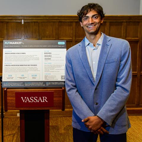 A person in a suit jacket stands next to a podium labeled "Vassar" on which a poster filled with writing pertaining to a business proposal is displayed.
