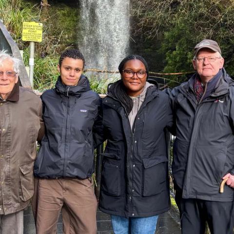 Four people stand outdoors, with foliage and a stone staircase behind them.