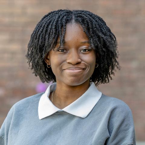 Portrait of a person smiling at the camera outdoors, wearing a light blue sweater with a white collared shirt. A brick wall appears softly blurred in the background.