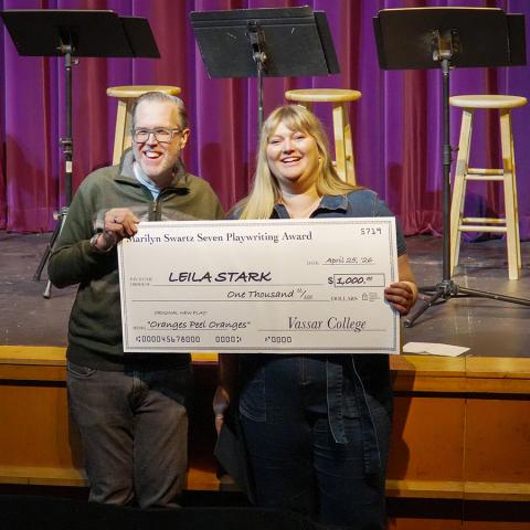 Two smiling people sit on a stage holding a replica of a giant check made out for $1,000.