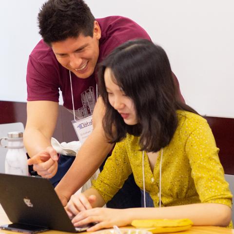 Two conference participants collaborate at a table, with one person pointing at a laptop screen while the other works on the computer. Both wear conference name badges, and reusable water bottles and laptops are visible in the bright classroom setting, suggesting a hands-on workshop or collaborative learning session.