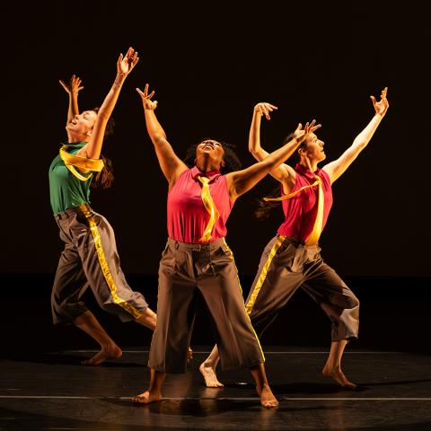 Three spotlit dancers strike poses with arms raised during a performance on a darkened stage.