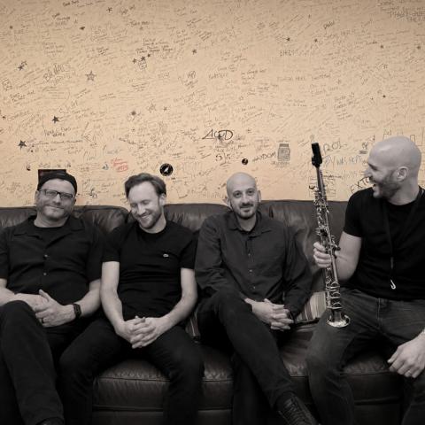 Four members of the True North Jazz Project sit on a couch in front of a graffiti-covered wall, with one holding a soprano saxophone.