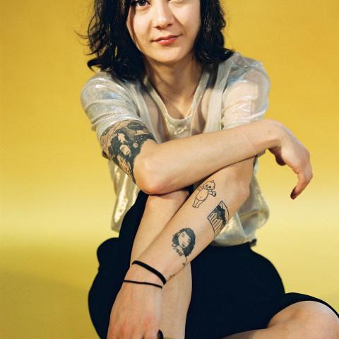 Michelle Zauner seated against a yellow background, wearing a sheer top and black skirt, with several tattoos visible on their arms.
