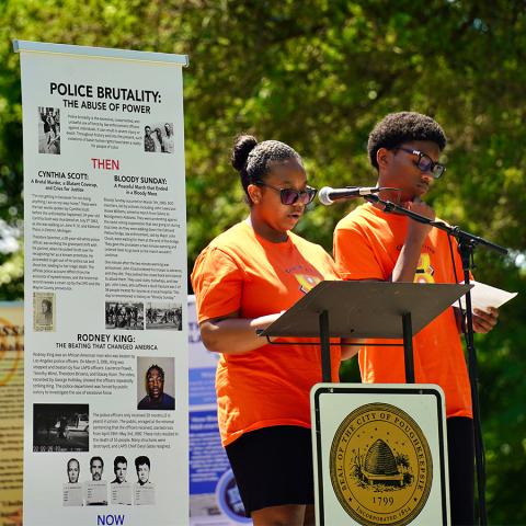 Two people standing at an outdoor podium next to a large poster about the history of police brutality.