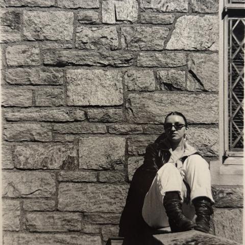 An artistic sepia-toned image of a person wearing sunglasses sitting in front of a stone wall.