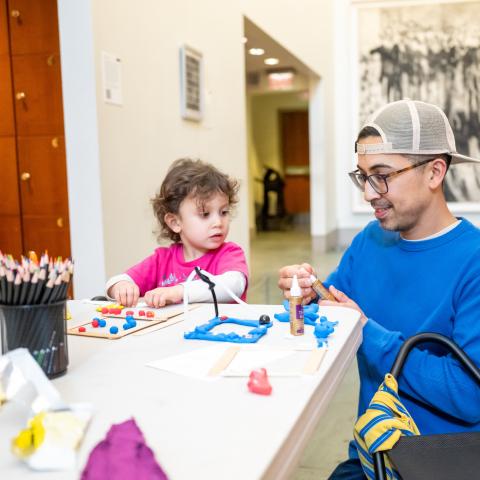 Photo of a man and young girl seated at a table making art with clay, colored pencils, and glue.