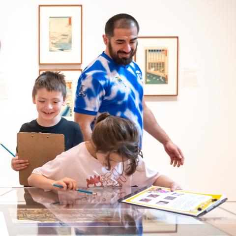 Photo of a man with two children in an art gallery. The children are holding clipboards and pencils, and looking at art in a case to do a scavenger hunt.