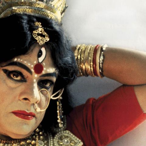 A person wears elaborate gold jewelry, a crown, red clothing, and theatrical facial makeup with a third eye, resting an arm stacked with bangles near their head.