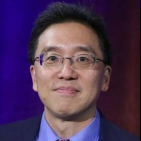Charles Tien wearing wire-rimmed spectacles and has short black hair and a slight smile, dressed in a dark suit jacket over a light blue collared shirt. The background is a solid, dark purple-blue color.