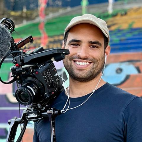 A smiling man stands beside a video camera and microphone, with a colorful mural behind him.