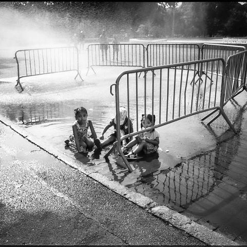 A black-and-white photograph taken by the artist Jas Leon. In the picture are three children playing in a spray of water while sitting in a city street.   